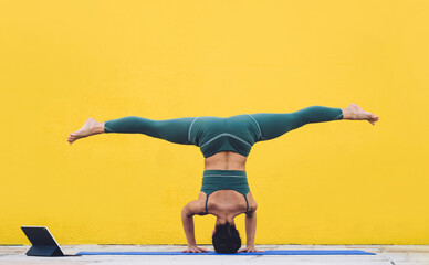 Slim female practicing yoga in tripod headstand and spiral legs pose using tablet in yard