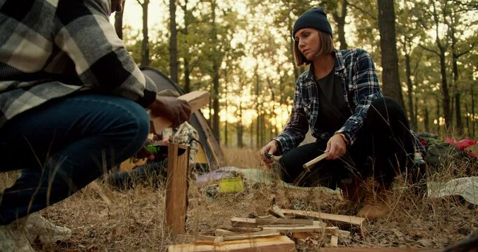 A girl in a black hat with a bob hairstyle helps a man with Black skin in a checkered shirt chop wood with a hunting knife during a camping stop in a sunny summer green forest