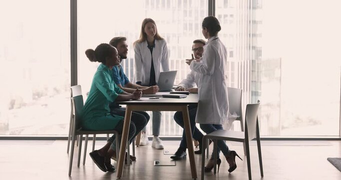 Discussion of multiethnic clinic staff gathered together in skyscraper office conference room. Five young medical workers take part in concilium, share patient disease prognosis, professional opinion