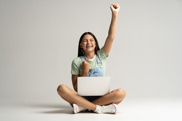 Happy smiling teen girl rejoycing sitting on gray background with laptop on legs raising hand up...