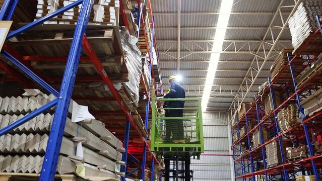 Team two male and female workers board forklift to inspect cardboard boxes on a pallet rack, secured by a team of safety drivers inside an export logistics manufacturing facility building.