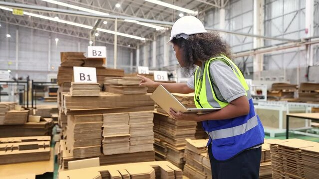 African American female worker standing checking pile of cardboard on pallet rack cardboard manufacturing industry plant packaging products logistics working recording data on laptop computer.