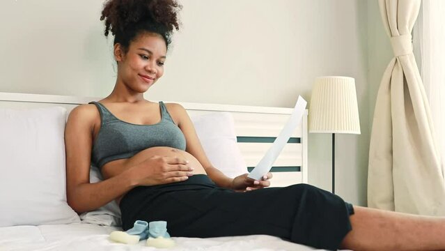 Happy portrait pregnant African American mother sitting gently rubbing her stomach on the bed looking at the ultrasound upcoming baby with anticipation and joy to see fetus's development healthy.