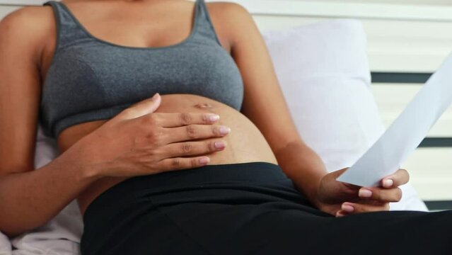 Happy portrait pregnant African American mother sitting gently rubbing her stomach on the bed looking at the ultrasound upcoming baby with anticipation and joy to see fetus's development healthy.