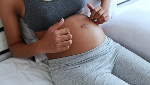 Pregnant African American woman sits on the bed and gently scratches her stomach with both hands. To relieve stomach itching caused by cracked skin, dry skin, and rashes.