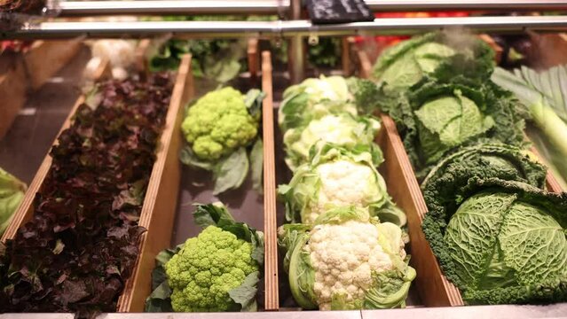 There are fresh vegetables on wooden shelf of the store. Different varieties of cabbage are laid out in separate containers on grocery store window