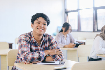 Smiling young man at college leaning on desk with classmates in background.