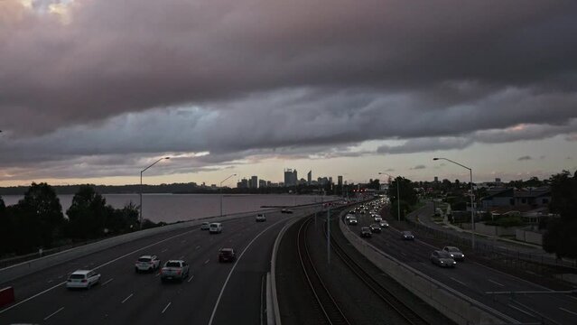 Grey Clouds With Purple Tinges Fly Overhead Above Wide Angle View Of Freeway Traffic At Dusk. Swan River, Perth City In Background. Trains Flash Past. Timelapse