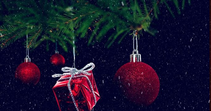 Animation of snow falling over hanging baubles and christmas gifts on a branch on black background