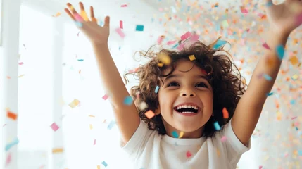 Fotobehang Geburtstag Konzept, Ein kleiner, fröhlicher Junge, umgeben von pastellfarbenem Konfetti, hebt an seiner Geburtstagsfeier glücklich lachend die Arme vor einem weißen Hintergrund © Jennifer