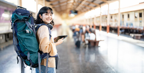 Young female traveler with backpack and using tablet, standing at train station. - obrazy, fototapety, plakaty