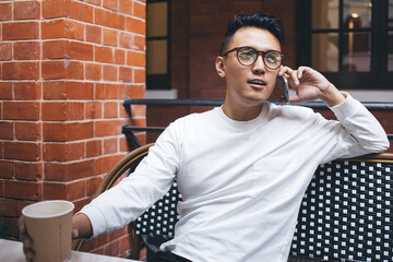 Concerned young Asian male talking on smartphone in cafe
