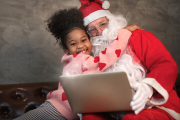 Little girl African hugging father Santa on couch at home, Santa places a laptop computer on his...