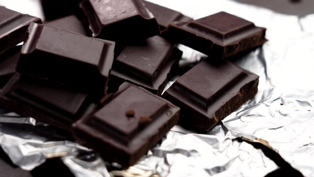 Broken crushed bar of bitter dark chocolate with pieces on shiny foil wrapping close up. Rotation