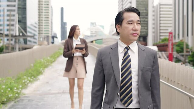 4K Confidence Asian businessman and businesswoman colleague holding briefcase and digital tablet walking city street at office district. Business people company employee go to work in the morning.