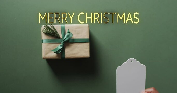 Merry christmas text in gold with gift and african amrican male hand holding blank tag on green