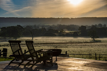Fototapeta na wymiar Reclining chairs, sit, and relax at a beautiful countryside view of a glorious sunrise over grassy rural landscape in Devon UK. Cattle grazing in the distance with a mist hanging over the fields. 
