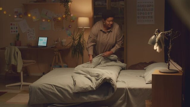 Caring African American grandma wearing long nightie putting her two beloved grandkids to sleep at night, wishing them goodnight and kissing on forehead