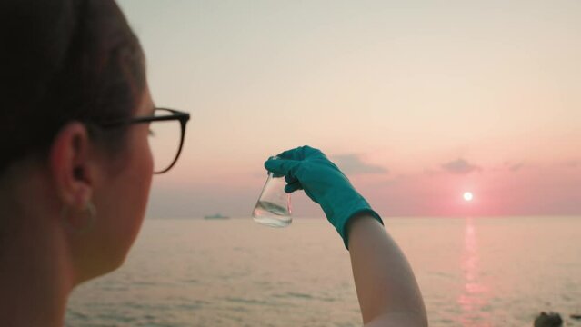 Back view of ecologist in rubber gloves shakes flask with sample of water from ocean. Sunset and sea are in background. Concept of ecology and chemical test.
