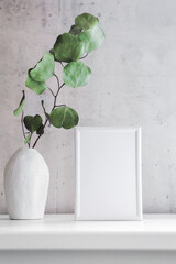 Ein leerer weißer Bilderrahmen und ein Vase mit einem Eukalyptuszweig auf einer Kommodes. Inneneinrichtung.
