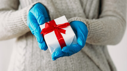 Christmas present. Winter gift. Unrecognizable human hands in protective gloves holding wrapped box distancing congratulating isolated on gray background.