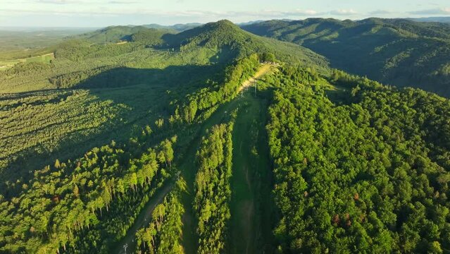 the drone on the right flies around the top of the mountain on which the ski slopes and lifts are located and opens a view of the meadow below the mountain. Summer, evening. Ukrainian Carpathians, 
