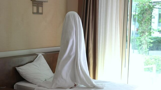 kid child jumping on bed with bed sheet blanket on head, imitating a halloween ghost 4k