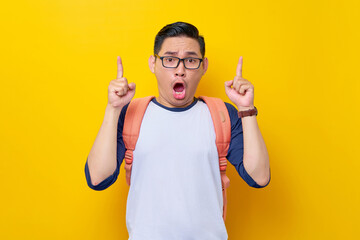 Surprised young Asian student man in casual clothes and glasses backpack pointing finger up at...