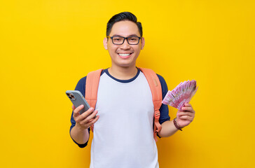 Smiling young Asian student man in casual clothes and glasses backpack holding  mobile phone and cash money isolated on yellow background. Education in High School University College concept