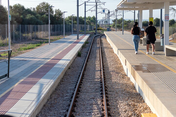 Plataforma de la estaci&oacute;n de tren con banco de piedra y valla. &Aacute;rboles y cielo azul con dos personas, no reconocibles, de espaldas caminando por la estaci&oacute;n direcci&oacute;n al infinito.