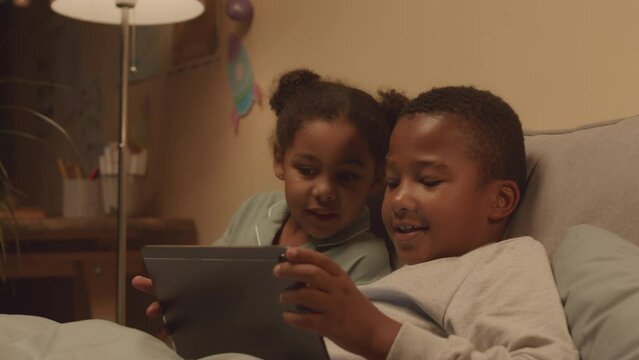 Waist up of African American little boy in girl playing games on tablet pc while lying together in soft warm bed at night