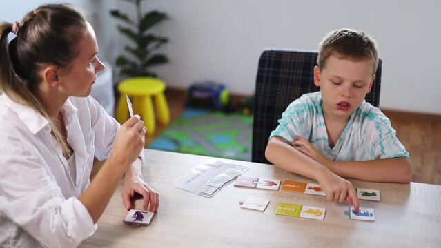 ABA Therapist Leads Therapy Session with Autistic Boy Using Color Cards; Rewards with Tokens; Bright Room with White Walls, Wooden Floor; Fiddle Leaf Fig (Ficus Lyrata) & Toys in the Background