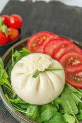 Sliced Italian cheese burrata is served on a round plate with basil and tomatoes with a fork and knife on the background of an old tree. Top view