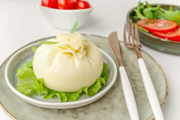 Italian soft burrata cheese is served on a round plate with basil and tomatoes with a fork and knife on a light background