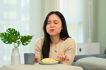Happy young asian woman sitting in living room and eating tasty pie with closed eyes. People, food and lifestyle