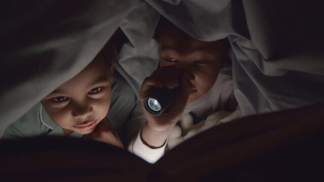 Medium closeup of two elementary age African American brother and sister using flash light while lying under warm duvet and reading fairy tales at night