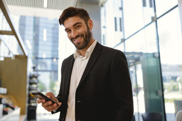 Male manager holding mobile phone and looking at camera with smile standing on office background