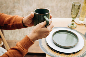 Men's hands in a warm terracotta hoodie hold a cup of coffee or tea in front of a panoramic window and breakfast table. Morning coffee, autumn breakfast, without a face