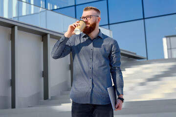 A well-groomed European man with a red beard and glasses checks his email using a tablet on the street