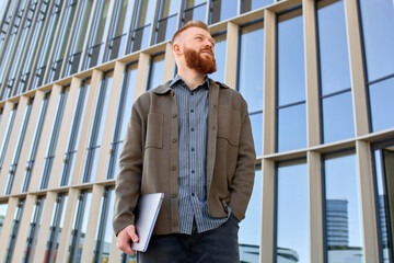 A handsome red-haired freelancer man with a beard stands confidently against the backdrop of a city building and looks into the distance