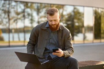 Fototapeta premium A 35-year-old man with a red beard sits on a bench with a laptop during lunch break