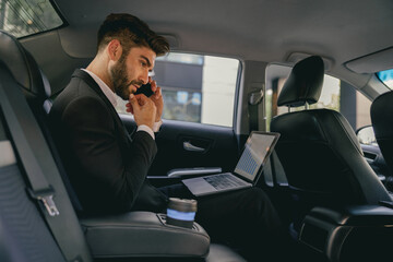 Handsome businessman in suit using personal computer and making phone call in taxi