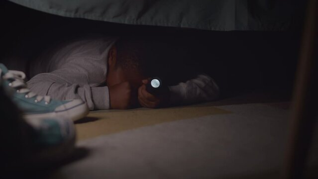 Little African American boy afraid of monsters lying under his bed with flashlight in dark room with creepy flickering light