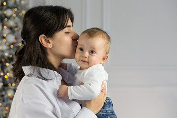 A young mother kisses her little child while standing near a Christmas tree