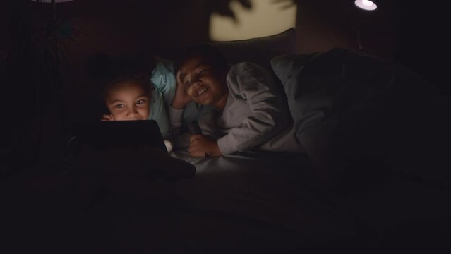 Two African American tween brother and sister using digital tablet while lying on bed in dark room with flashlight flickering and spooky blur shadows on wall playing game