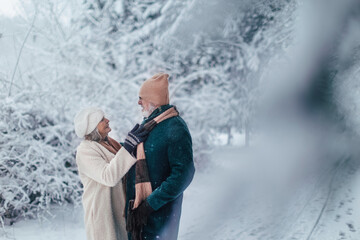 Elegant senior couple walking in the snowy park, during cold winter snowy day. Elderly woman fasten husband's winter coat. Wintry landscape.
