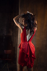 A femme fatale in a red dress. Attractive brunette in an elegant dress, with a cutout on the back. photo in dark colors on a textured wall, warm light