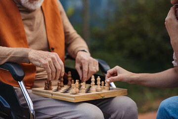 Senior man playing chess outdoors with his daughter. Nursing home client in wheelchair spending...