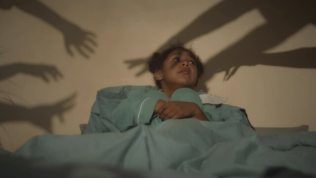 Slowmo of scared African American little girl sitting on bed in dark room and trying to make unrecognizable monsters leave. Spooky hand silhouettes playing shadows on wall trying to scare her