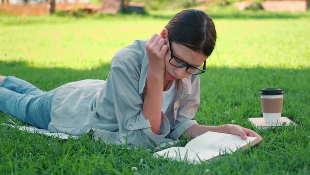 Teenage schoolgirl studying reading her books, lying down outdoors. Back to school, dreaming, inspiration. Student girl reads book on the green grass in the college yard or park. Distance learning 4K	